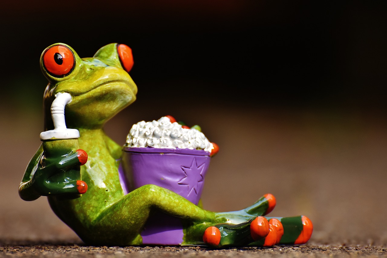 a cartoon frog with a drink in one hand and a tub of popcorn between its legs